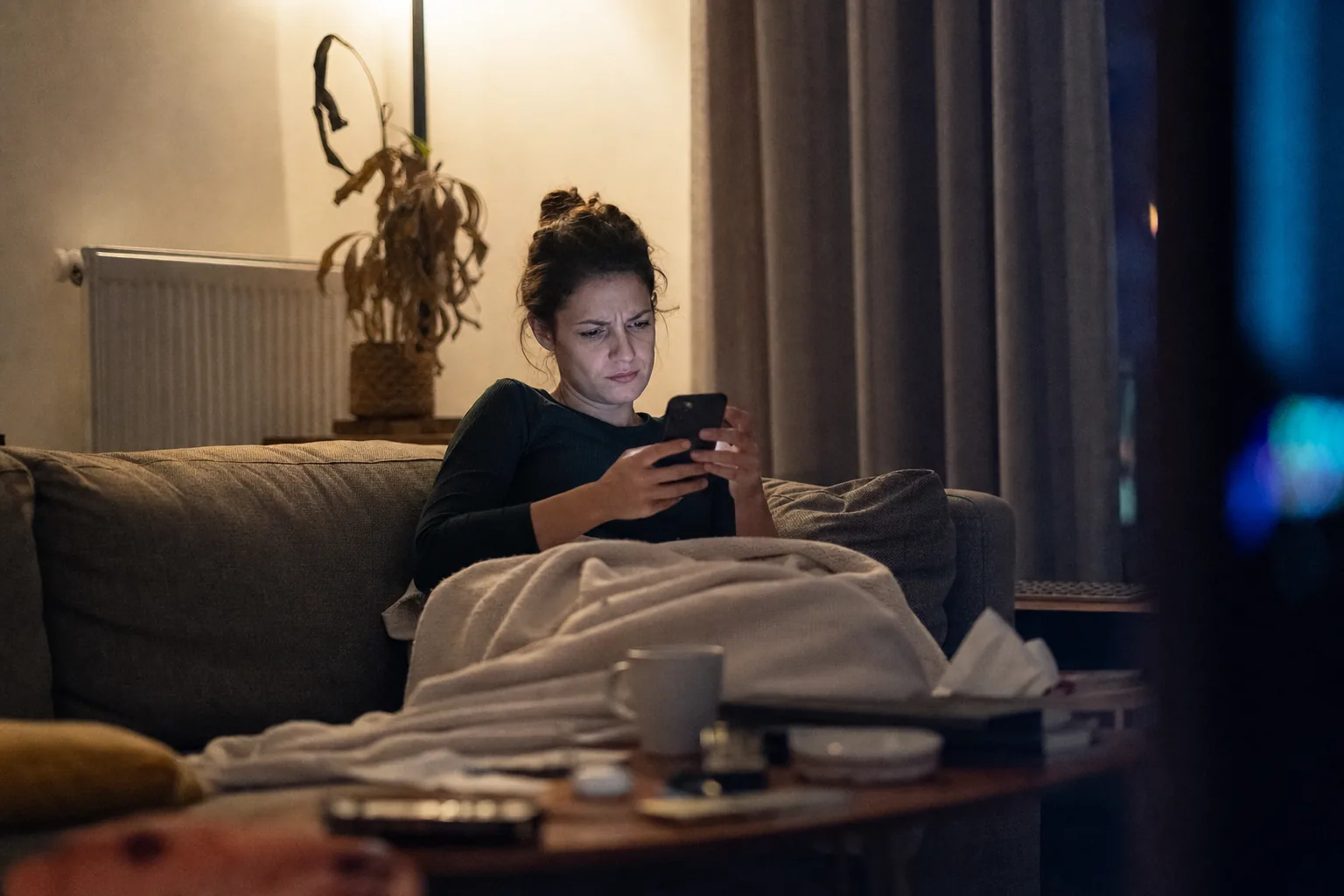Dans un salon éclairé par une seule lampe, une femme assise dans un canapé sous une couverture regarde son téléphone en fronçant les sourcils, une tasse et des objets posés sur la table basse