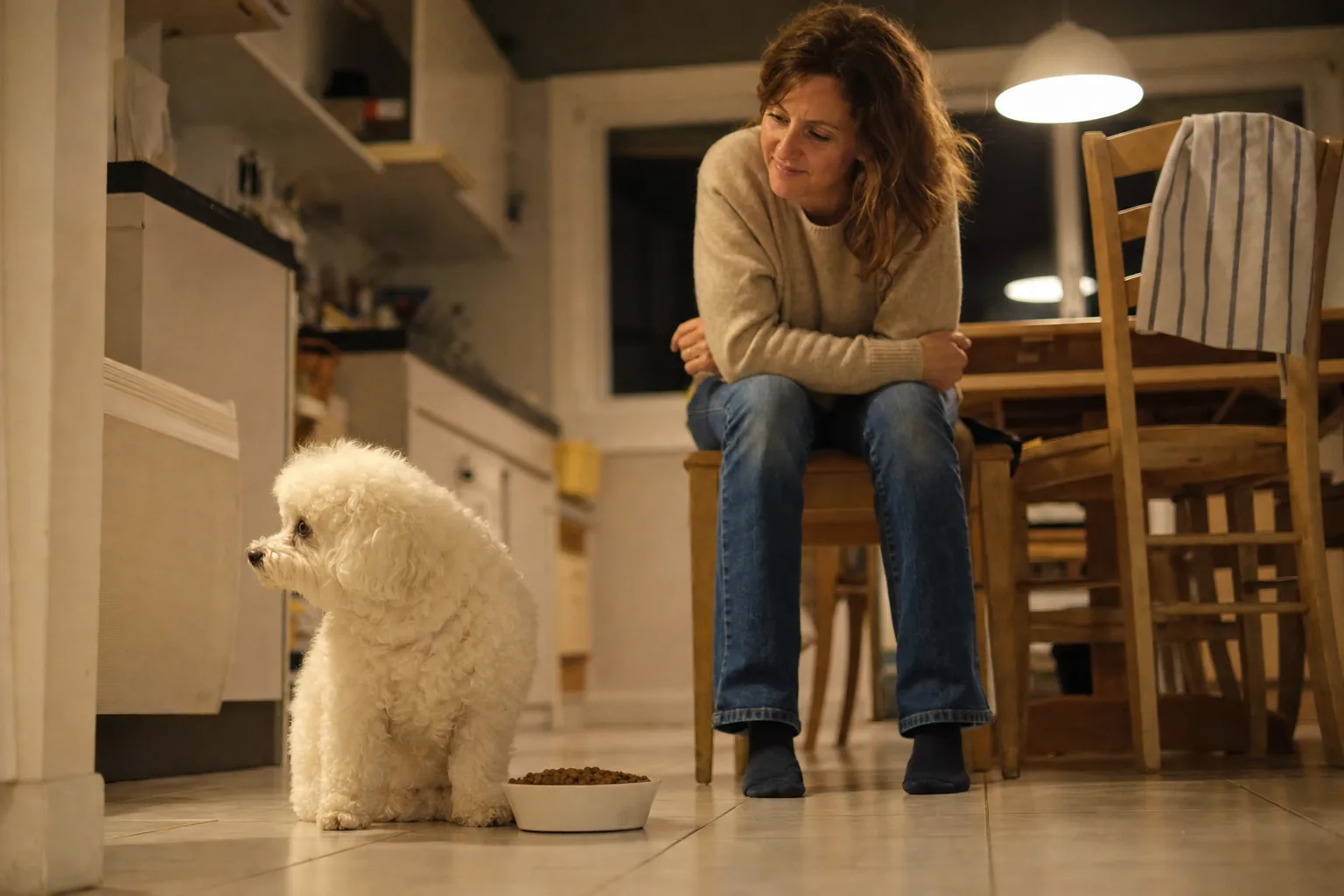 Le soir, dans la cuisine d’un appartement, un bichon frisé blanc assis à côté de sa gamelle de croquettes détourne la tête, sous le regard amusé d’une femme en pull beige assise sur une chaise, les coudes sur les genoux