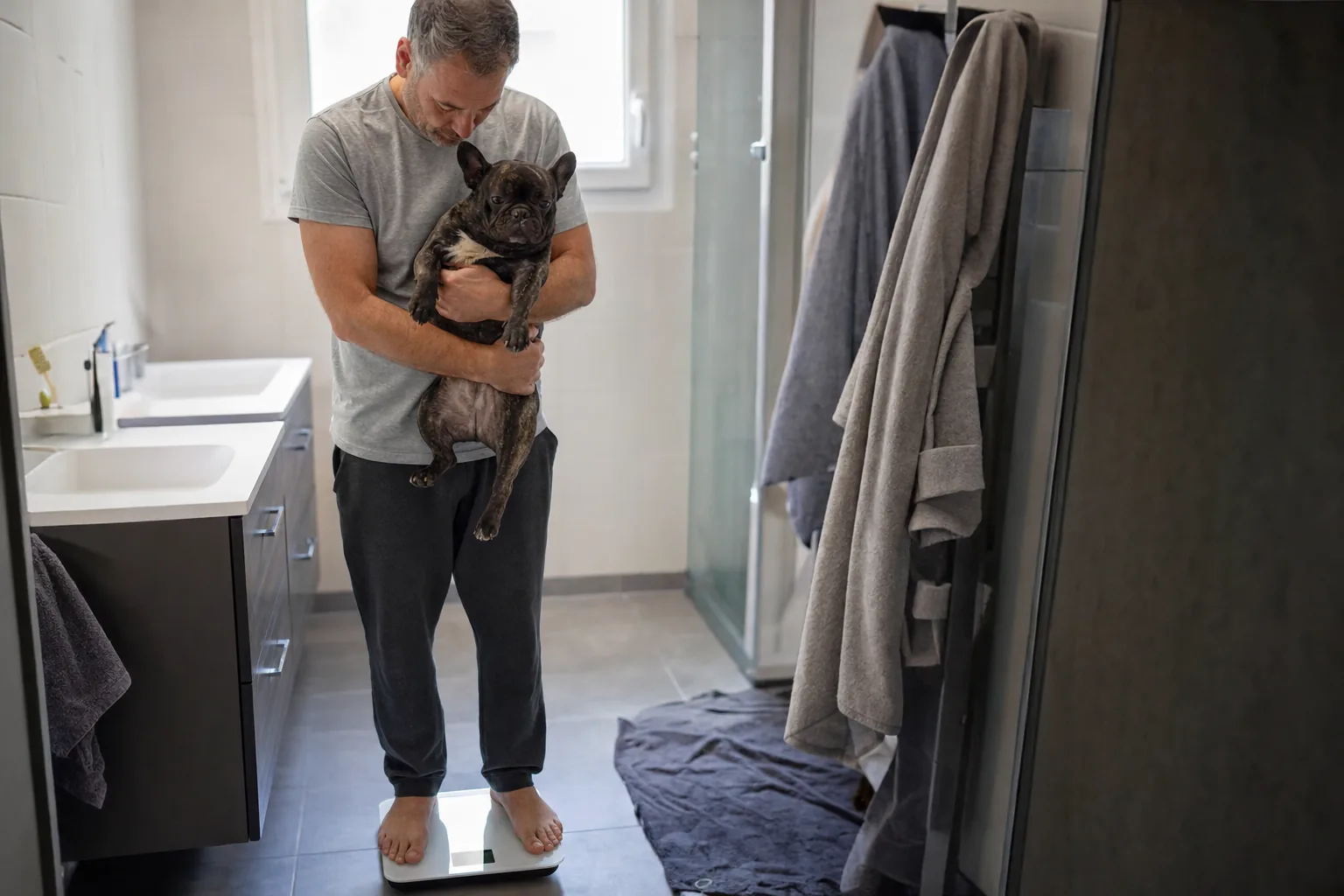 Dans une salle de bains, un homme en tee-shirt gris debout pieds nus sur un pèse-personne tient dans ses bras un bouledogue français bringé et baisse les yeux vers la balance
