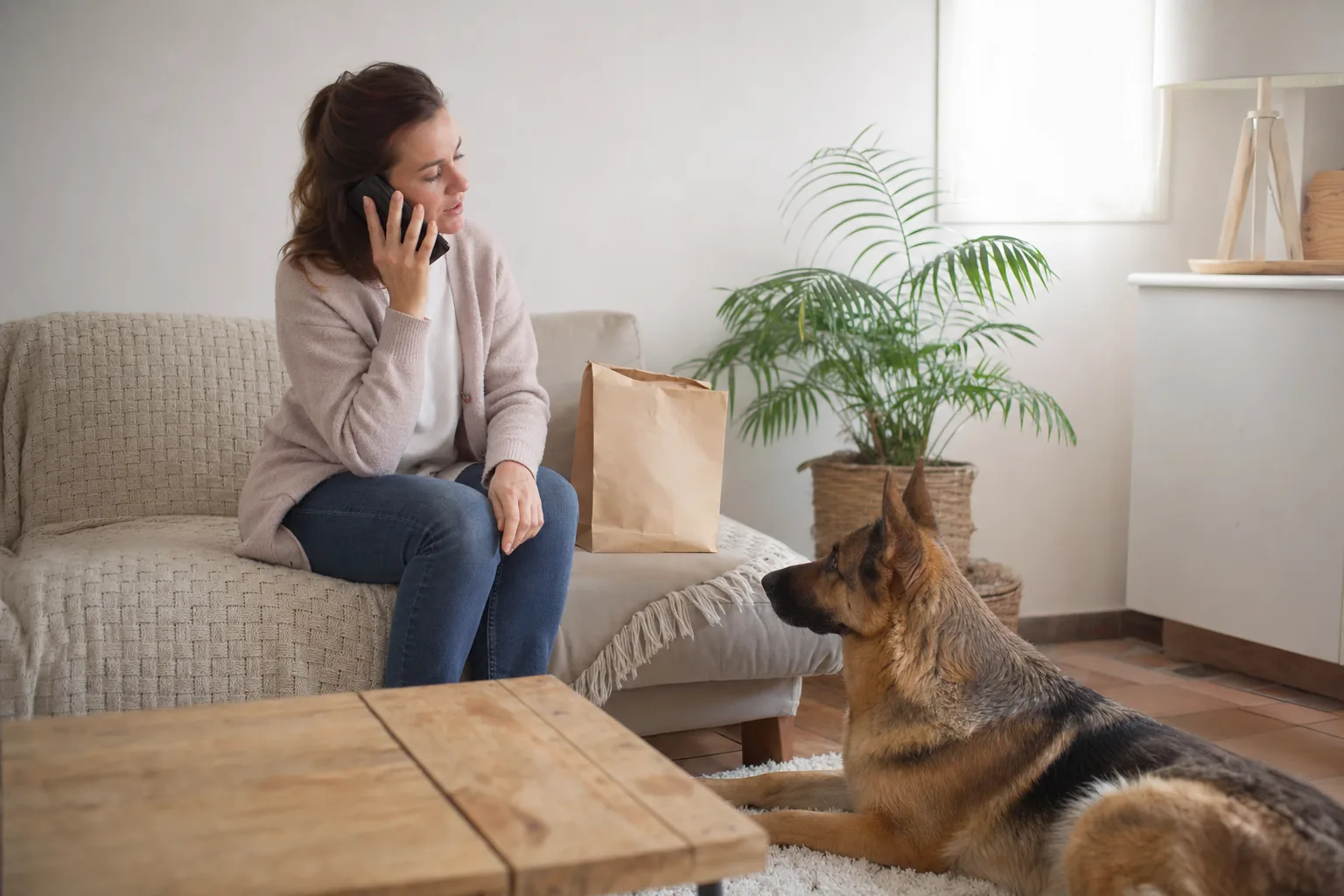 Femme assise sur un canapé qui téléphone, un sac en papier kraft posé à côté d’elle, un berger allemand couché sur le tapis qui la regarde