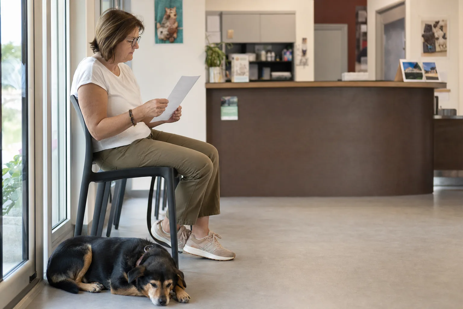 Dans la salle d’attente d’une clinique vétérinaire, une femme lit une feuille de devis pendant que son chien noir et feu dort à ses pieds