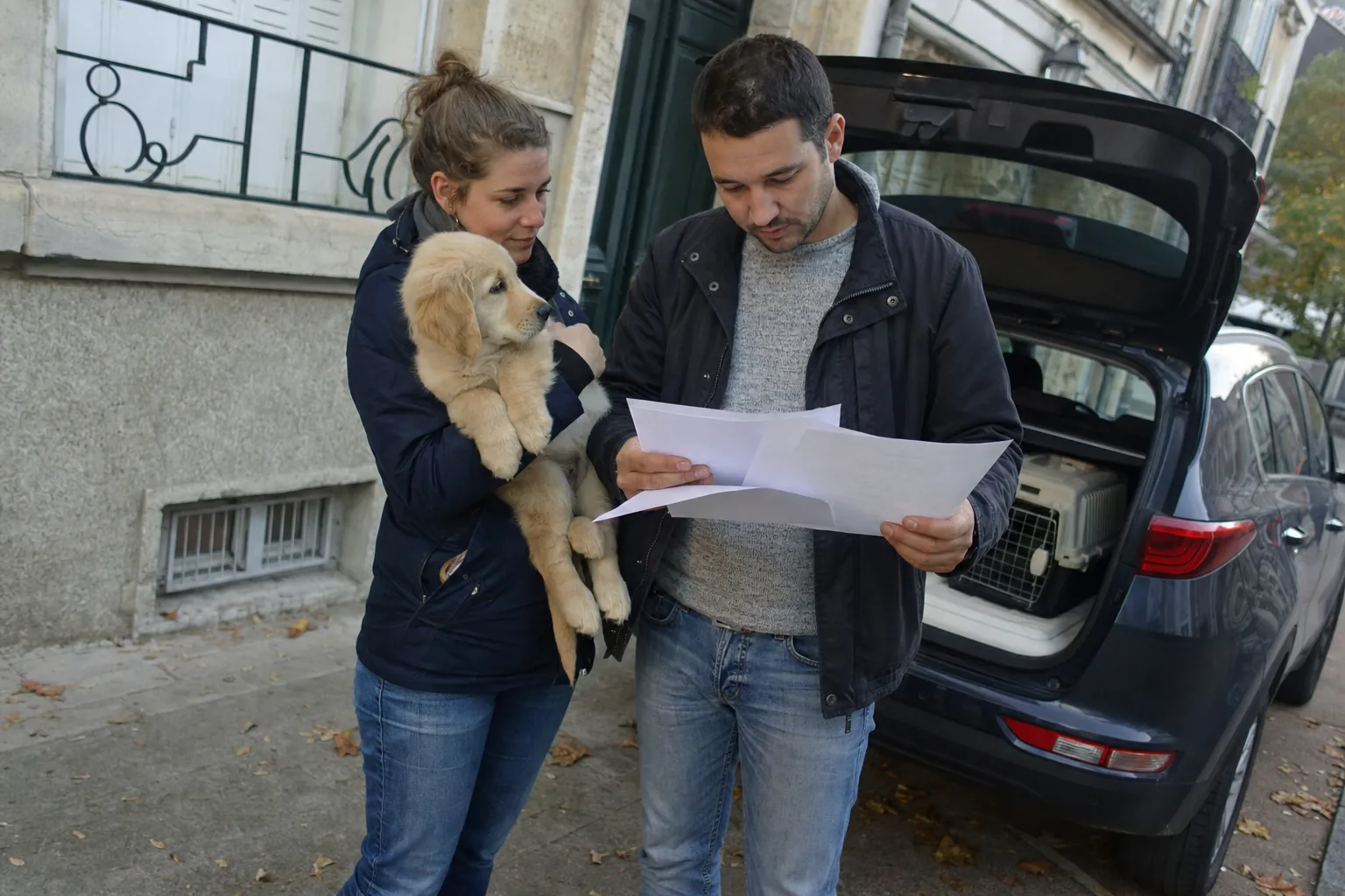 Sur un trottoir devant un immeuble ancien, une femme tient un chiot golden retriever dans les bras pendant qu’un homme lit des feuilles imprimées, une caisse de transport posée dans le coffre ouvert derrière eux