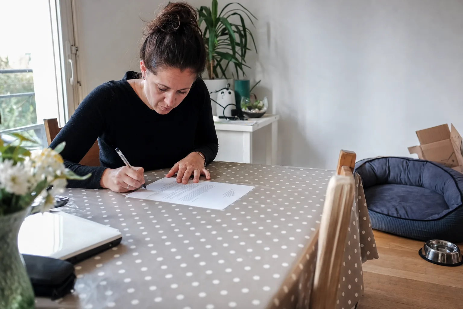 Une femme seule signe un document imprimé à la table de sa salle à manger, un panier pour chien encore vide et une gamelle en inox posés dans le coin de la pièce
