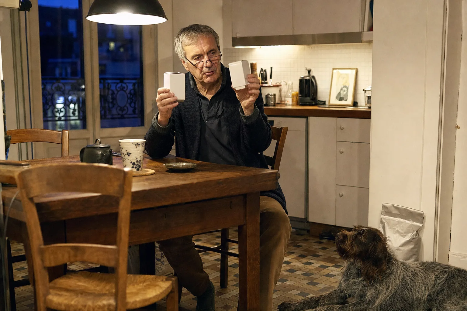 Homme âgé à lunettes assis à la table d’une cuisine parisienne, qui compare deux petites boîtes blanches sous la lampe, un griffon korthals couché près de lui