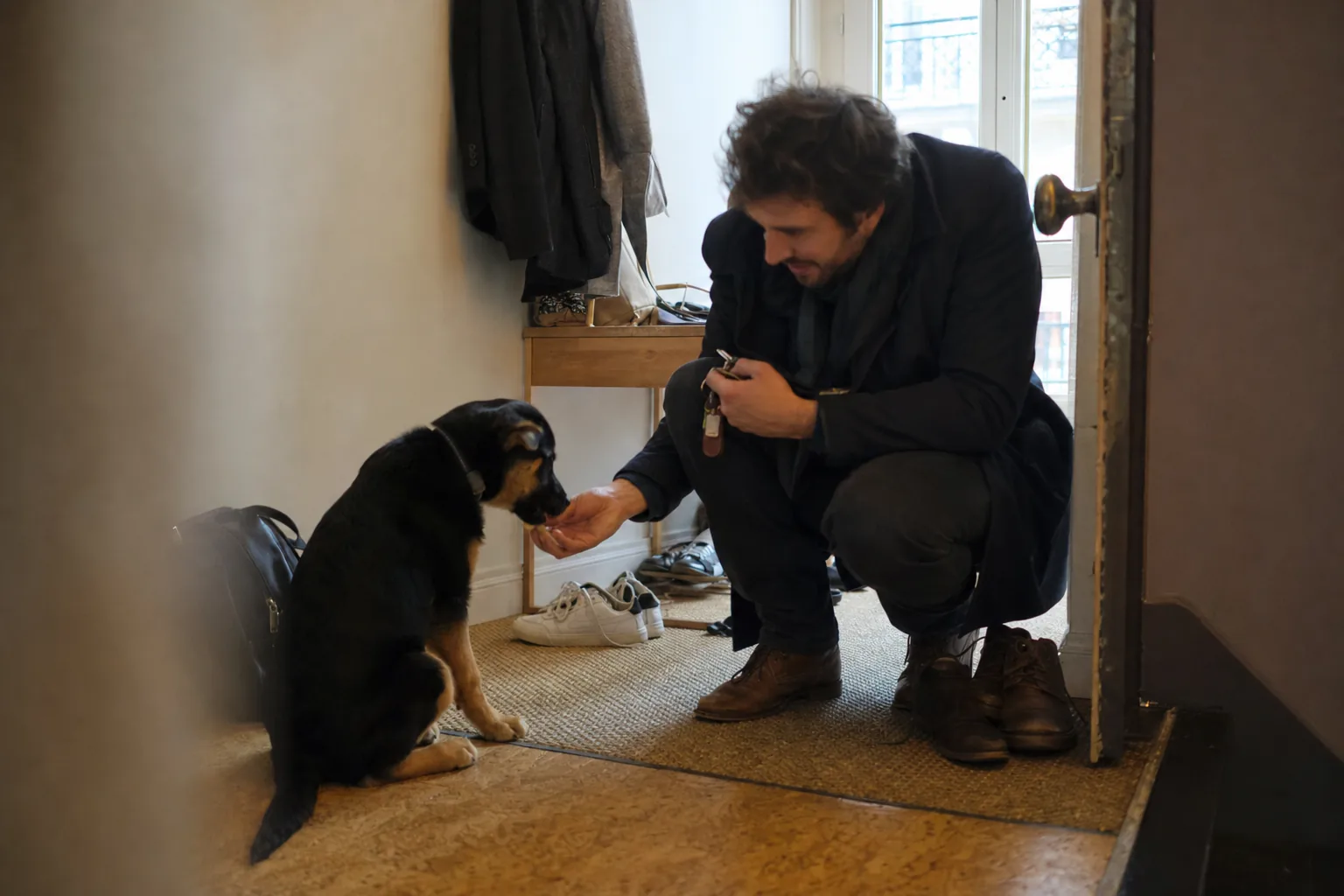 Dans l’entrée d’un appartement, un homme en manteau, les clés à la main, s’accroupit pour donner une friandise à un jeune chien croisé noir et feu assis près des chaussures, juste avant de sortir.